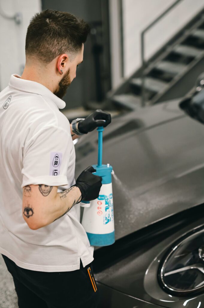 Un technicien expert de l'Atelier Clean UP, portant un polo blanc et des gants noirs, utilise un pulvérisateur manuel pour appliquer de la mousse active sur le capot gris d'une voiture dans son atelier à Metz.